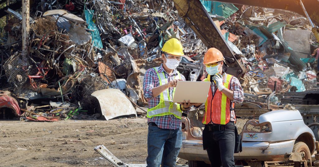 Two workers in safety gear and masks examine a laptop at a scrapyard with metal debris in the background.